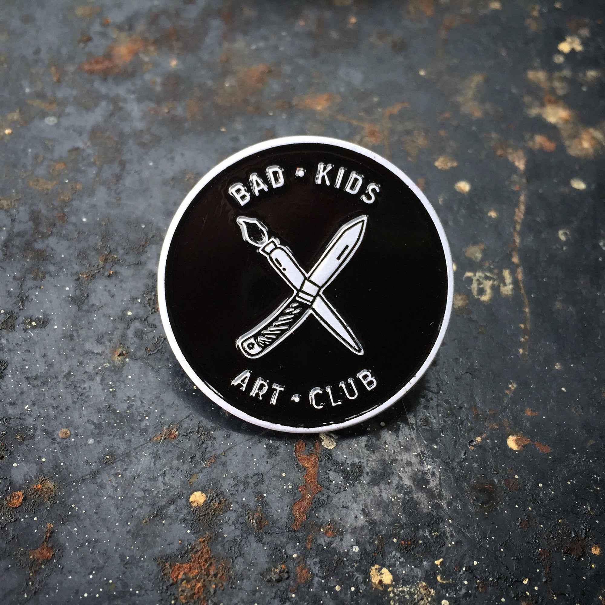 Circle enamel pin with "x" of paintbrush and knife with Bad Kids Art Club around it. By Print Ritual
