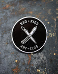 Circle enamel pin with "x" of paintbrush and knife with Bad Kids Art Club around it. By Print Ritual