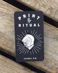Black square enamel pin of skeletal hand leaving print on bare bottom. Image in white. On Print Ritual Card.